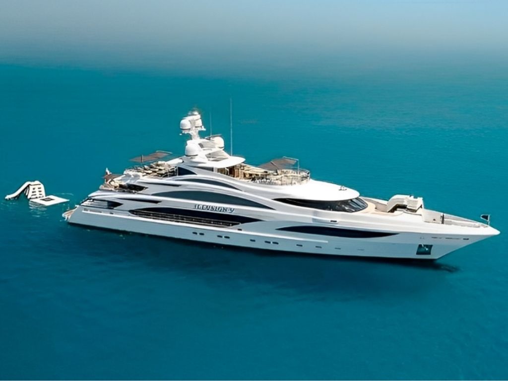 An aerial view of an 88 Sirena yacht with its tender in tow, cruising through vibrant turquoise Caribbean waters past a secluded island anchorage at sunrise.
